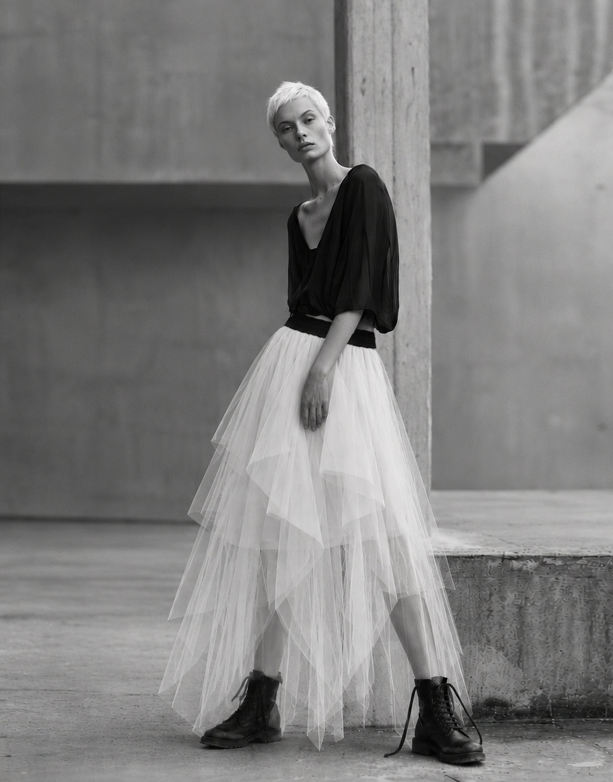 A model with short platinum blonde hair stands in an urban concrete setting, wearing a black asymmetrical top, a long layered white tulle skirt, and black combat boots.