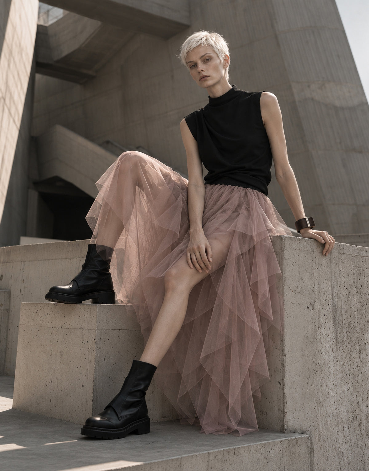 A model with short blonde hair sits on concrete steps wearing a black sleeveless mock-neck top, a long dusty pink tulle skirt, and black leather ankle boots.