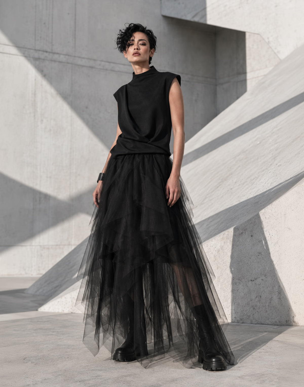 A model wearing a black sleeveless mock-neck top and a long, layered black tulle skirt, standing against a minimalist concrete architectural background with sharp geometric shadows.