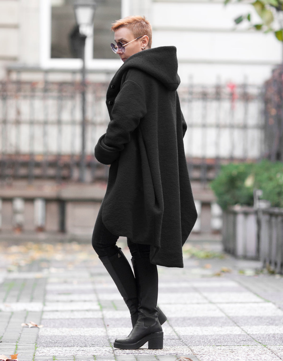 A person standing outdoors on a paved walkway, wearing a long black hooded coat.